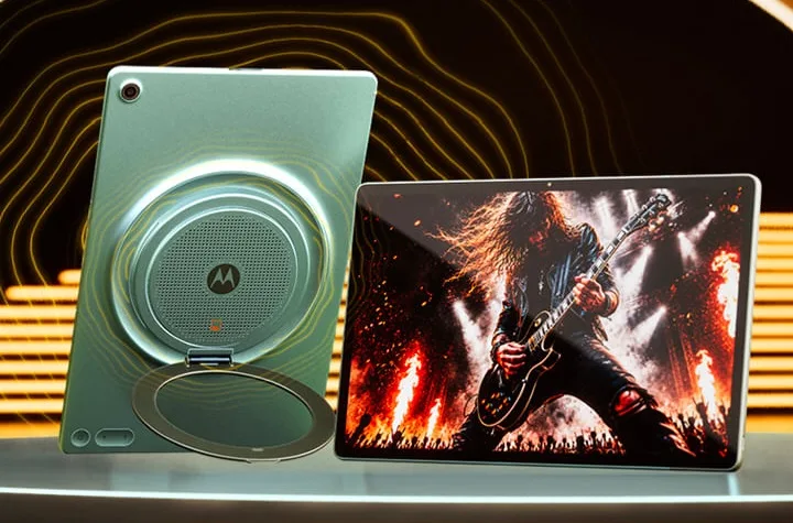 Product image of the Moto Pad 70 Groove, showing both the back and front of the tablet. On the left, the light green (Pantone Blue Surf) rear panel of the tablet stands upright using a built-in circular ring kickstand. The center of the back panel features a prominent, circular metallic speaker grille with the Motorola logo and a small orange JBL logo inside it. On the right, the front display is shown landscape-style, displaying a high-contrast image of a guitarist playing on a stage with fire effects.