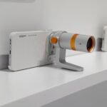 Oppo Find X10 Launched in China with Dual 200MP Cameras, Dimensity 9600M, 1.5K AMOLED Display, and 8,000mAh Battery 1 Oppo Find X10 with Hasselblad telephoto lens accessory attached, white textured case with orange adjustment knobs, displayed on white shelf