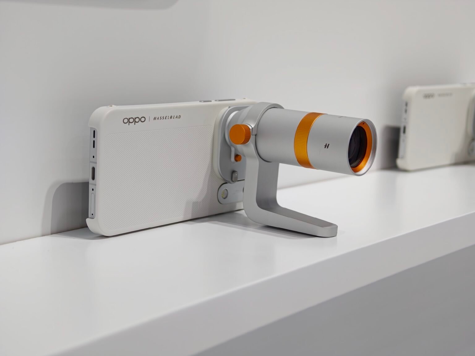 Oppo Find X10 with Hasselblad telephoto lens accessory attached, white textured case with orange adjustment knobs, displayed on white shelf
