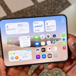 iPhone Duo foldable smartphone held open showing large inner display with iOS home screen widgets and app icons, crisp detailed official look