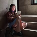 Young woman seated on concrete stairs using the starlight‑finish iPad mini 8 (2026) beside a fabric bag, with textured concrete walls and a window in the background.