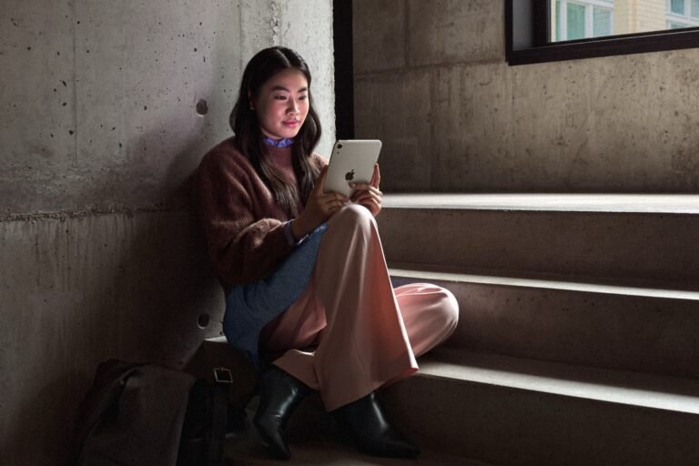 Young woman seated on concrete stairs using the starlight‑finish iPad mini 8 (2026) beside a fabric bag, with textured concrete walls and a window in the background.